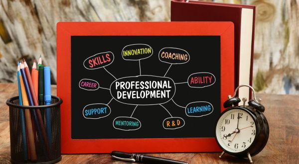image of a desktop with a pencil holder with pencils on the left, a chalkboard with a pen at it's base in the middle and a clock to the right of that. chalkboard has a word map for professional development. words written are: innovation, coaching, ability, learning, r and d, mentoring, support, career, and skills 